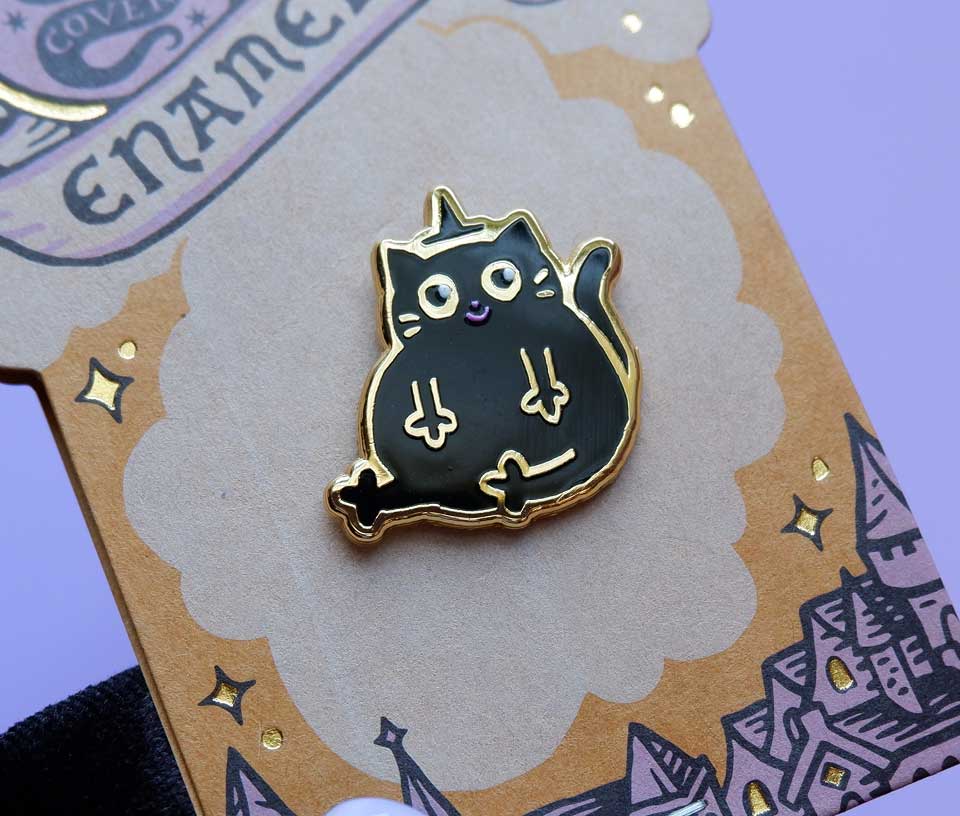 A black cat enamel pin with gold metal outlines and small purple and white details, displayed on a decorative brown card against a lavender background.