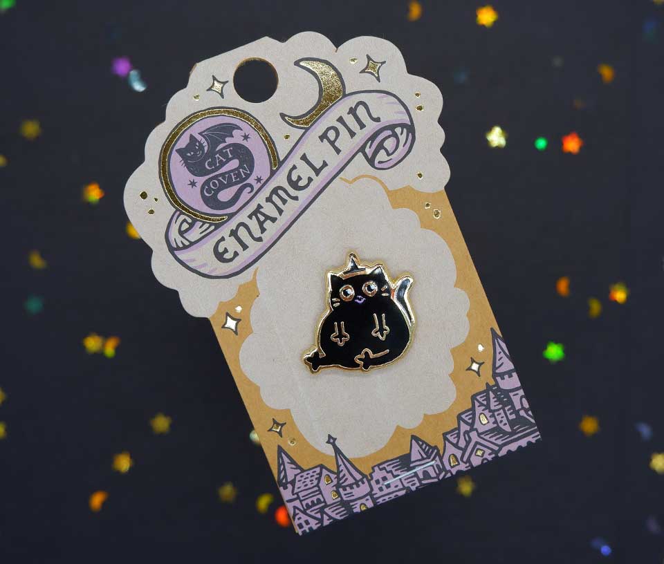 A black cat enamel pin with gold details and a crescent moon accessory, mounted on a decorative illustrated backing card that reads 'ENAMEL PIN' against a dark glittery background.